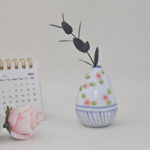 May include: A white ceramic vase with a floral pattern of pink and green dots, and blue stripes at the base. A calendar showing October 2025 and two pink roses are in the foreground. A dark grey plant stem is in the vase.