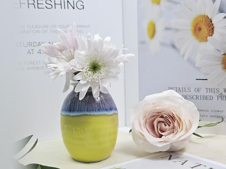 Puede incluir: Un peque&ntilde;o jarr&oacute;n de cer&aacute;mica amarillo y azul con un arreglo de flores blancas y rosas. El jarr&oacute;n tiene una forma redondeada y una superficie texturizada. Las flores est&aacute;n dispuestas en un estilo simple y elegante.