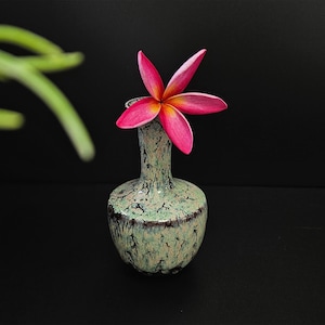May include: A single pink flower with a yellow centre in a small, green and black ceramic vase.