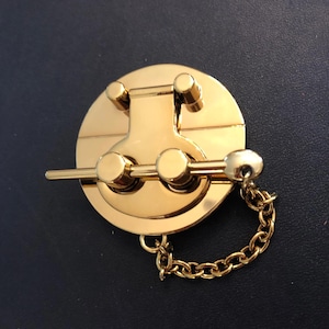 May include: A gold-colored metal clasp with a circular base and a rectangular top. The clasp has a chain attached to it. The background is a dark blue color.