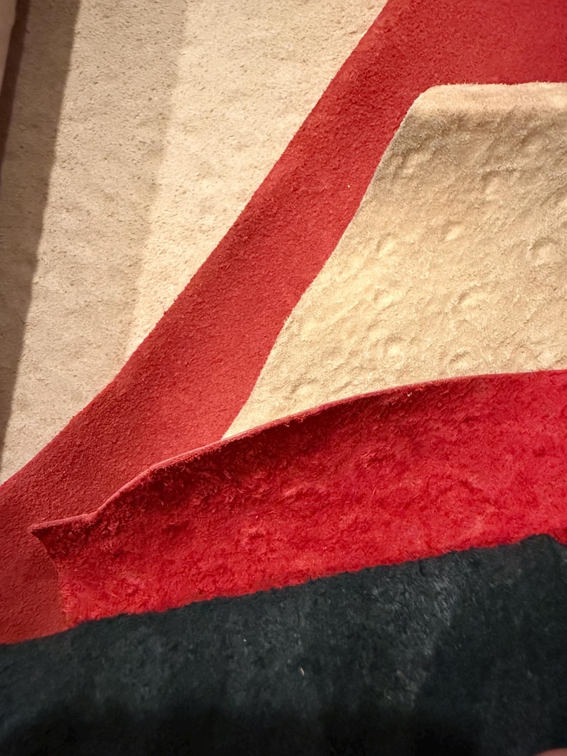 Peut inclure: Gros plan sur un tapis textur&eacute; avec un motif compos&eacute; de sections rouges, beiges et bleu fonc&eacute;. La section rouge forme une bande diagonale sur le fond beige, avec une section bleu fonc&eacute; en bas.