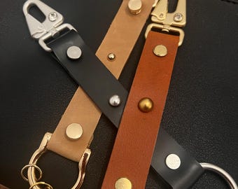 May include: Three leather keychains with different colored straps and metal clasps. The straps are brown, tan, and black. The clasps are silver and gold.