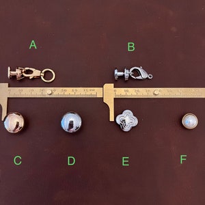 May include: Six metal hardware pieces for jewelry making, including two clasps, a round gold-toned stud, a round silver-toned stud, a silver-toned clover-shaped stud, and a white pearl-like stud. The hardware is displayed on a brown leather surface with a ruler for scale.