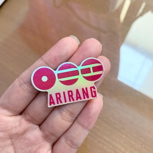 Pu&ograve; includere: Un adesivo olografico con un design rosa e rosso. L'adesivo presenta tre cerchi collegati con linee orizzontali e la parola "ARIRANG" in grassetto, in rosa.