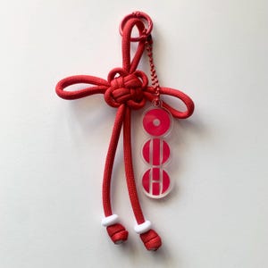 Puede incluir: Un llavero rojo con un diseño anudado y una pequeña cadena. El llavero tiene un dije de acrílico transparente con las letras "OEH" en rojo. Los extremos del llavero tienen detalles blancos.