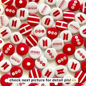 May include: A collection of circular pin-back buttons featuring red and white designs. Some buttons display the text ARIRANG BTS WORLD TOUR, while others feature red and white geometric logos, horizontal stripes, or circular patterns on white backgrounds.