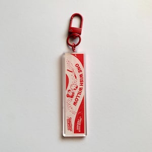May include: A rectangular acrylic keyring with a red lobster clasp. The insert features a cream background with red text reading ONE WISH WILLOW, CRACK, AMAZE YOUR FRIENDS, and You only get ONE WISH, accompanied by small red star and character graphics.