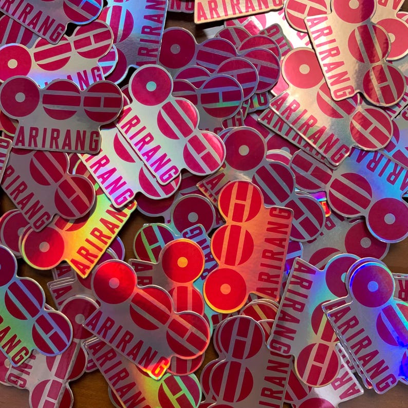 Pu&ograve; includere: Un mucchio di adesivi olografici con un design rosa e bianco. Ogni adesivo presenta la parola "ARIRANG" e una "H" stilizzata all'interno di una forma circolare e rettangolare. Gli adesivi hanno una finitura lucida e iridescente.