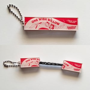 May include: A rectangular acrylic keyring with a silver ball chain, featuring red and white graphics with the text ONE WISH WILLOW, AMAZE YOUR FRIENDS, and You only get ONE WISH. The keyring splits into two pieces connected by a black cord.