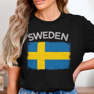 Könnte beinhalten: Schwarzes T-Shirt mit dem Wort "SWEDEN" in Weiß, über einem verwitterten schwedischen Flaggen-Design. Die Flagge hat ein gelbes Kreuz auf blauem Grund. Ein lässiges T-Shirt für Schweden-Liebhaber.