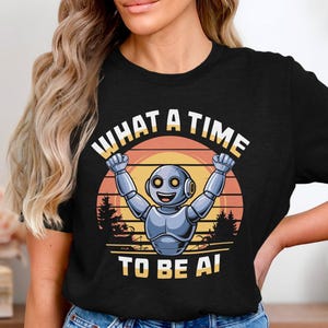Könnte beinhalten: Schwarzes T-Shirt mit einem Cartoon-Roboter, der die Arme zum Feiern hebt. Der Roboter ist vor einem Sonnenuntergangshintergrund platziert, mit dem Text "WHAT A TIME TO BE AI" in einem Kreis.