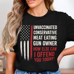 Könnte beinhalten: Schwarzes T-Shirt mit einem verblassten amerikanischen Flaggen-Design auf der linken Seite. Der Text lautet: "UNVACCINATED CONSERVATIVE MEAT EATING GUN OWNER HOW ELSE CAN I OFFEND YOU TODAY?" in Weiß und Rot.