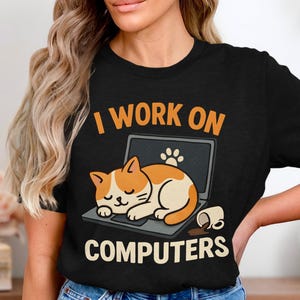 Könnte beinhalten: Schwarzes T-Shirt mit dem Text "I WORK ON COMPUTERS". Das Shirt zeigt eine Cartoon-Katze, die auf einem Laptop schläft, mit einem Pfotenabdruck auf dem Bildschirm und einer verschütteten Kaffeetasse.