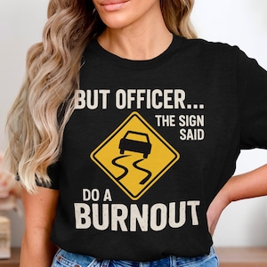 Könnte beinhalten: Schwarzes T-Shirt mit dem Text "BUT OFFICER... THE SIGN SAID DO A BURNOUT". Das Design beinhaltet ein gelbes Raute-Verkehrsschild mit einem schleudernden Auto. Der Text ist in weißer, fetter Schrift.
