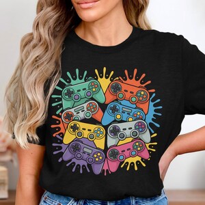 Könnte beinhalten: Schwarzes T-Shirt mit einer bunten Grafik von mehreren Videospiel-Controllern. Die Controller sind in verschiedenen Farben, darunter Grün, Orange, Blau, Gelb, Lila und Pink, mit einem Spritzer-Design. Ein lustiges Design für Gamer.