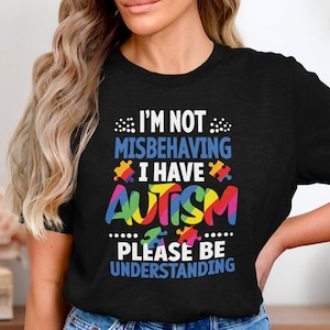 Puede incluir: Camiseta negra con el texto "I'M NOT MISBEHAVING I HAVE AUTISM PLEASE BE UNDERSTANDING". La palabra "AUTISM" está en colores arcoíris. El diseño incluye piezas de rompecabezas.