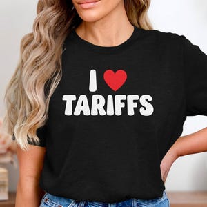 Könnte beinhalten: Schwarzes T-Shirt mit dem Text "I ❤️ TARIFFS" in Weiß, mit einem roten Herzsymbol. Das Shirt hat einen Rundhalsausschnitt.