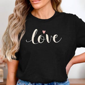 Könnte beinhalten: Ein schwarzes T-Shirt mit dem Wort "love" in weißer Kursivschrift und einem kleinen rosa Herz über dem Wort.