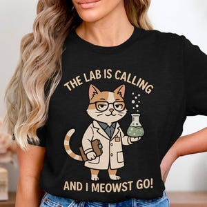 Könnte beinhalten: Schwarzes T-Shirt mit einer Cartoon-Katze im Laborkittel, die einen Kolben hält. Die Katze trägt eine Brille und der Text lautet "THE LAB IS CALLING AND I MEOWST GO!" Die Katze ist braun und weiß. Der Kolben enthält eine grüne Flüssigkeit.