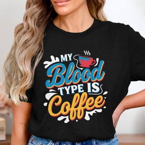 Könnte beinhalten: Schwarzes T-Shirt mit einem grafischen Design, auf dem in einer bunten Retro-Schrift "My Blood Type Is Coffee" steht. Oberhalb des Textes befindet sich eine kleine rote Kaffeetasse mit Dampf.