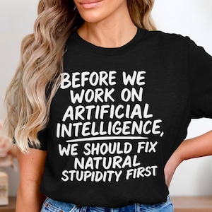 Op de afbeelding: Zwart T-shirt met witte tekst: "VOORDAT WE AAN KUNSTMATIGE INTELLIGENTIE WERKEN, MOETEN WE EERST NATUURLIJKE STUPIDITEIT OPLOSSEN." Ronde hals.