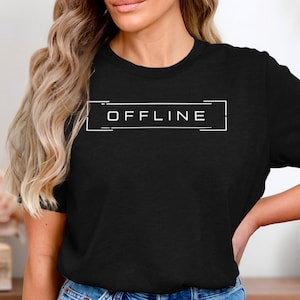 Könnte beinhalten: Schwarzes T-Shirt mit dem Wort "OFFLINE" in einem weißen rechteckigen Rahmen. Der Text ist mittig auf der Brust platziert. Das T-Shirt hat einen Rundhalsausschnitt.