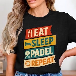 Könnte beinhalten: Schwarzes T-Shirt mit dem Text "EAT SLEEP PADEL REPEAT" in einem farbenfrohen Design. Das Design beinhaltet eine Gabel, ein Bett, einen Padel-Schläger und einen kreisförmigen Pfeil. Das Shirt ist ein Freizeitartikel.