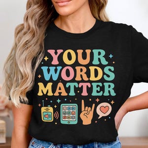 May include: Black t-shirt with the text "YOUR WORDS MATTER" in colorful retro font. The design includes illustrations of a retro phone, a tablet, a hand making a rock on gesture, and a heart-shaped speech bubble.