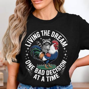 Könnte beinhalten: Schwarzes T-Shirt mit einem farbenfrohen Motiv eines Hahns und eines Waschbären. Das Design enthält den Text "LIVING THE DREAM ONE BAD DECISION AT A TIME" in kreisförmiger Anordnung. Der Hahn ist rot, blau und grün.
