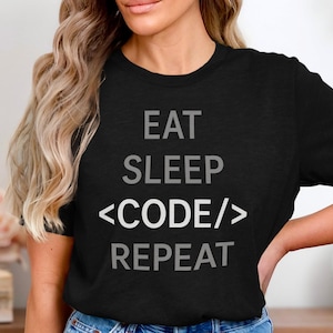 Könnte beinhalten: Schwarzes T-Shirt mit dem Text "EAT SLEEP <CODE/> REPEAT" in Grau und Weiß. Das Shirt hat einen Rundhalsausschnitt und ist aus einem weichen Material gefertigt. Das Shirt ist ein Freizeitartikel.
