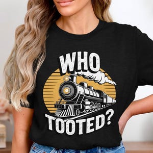 以下が含まれることがあります： 黄色い円の中に列車のグラフィックと白い文字「WHO TOOTED？」が入った黒いTシャツ。列車は白黒で、煙が出ています。Tシャツはクルーネックです。デザインは中央に配置されています。