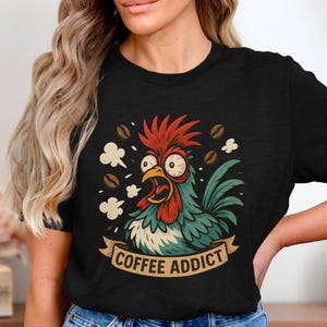 May include: Black t-shirt with a cartoon rooster design. The rooster has a red comb, blue feathers, and an open beak, surrounded by coffee beans and steam. A banner below reads "COFFEE ADDICT".
