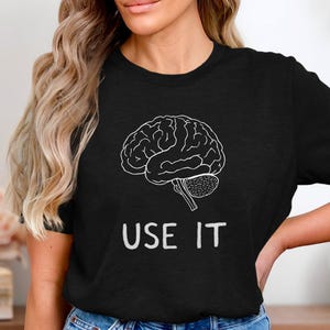 Peut inclure: Un t-shirt noir avec un contour blanc d'un cerveau et le texte "USE IT" imprimé sur le devant.