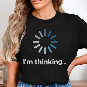 Puede incluir: Camiseta negra con un símbolo de carga en tonos azules y grises. El texto "I'm thinking..." está impreso debajo. La camiseta es de cuello redondo.
