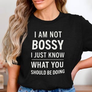 Könnte beinhalten: Schwarzes T-Shirt mit dem weißen Text "I AM NOT BOSSY I JUST KNOW WHAT YOU SHOULD BE DOING." Das Shirt hat einen Rundhalsausschnitt und ist aus weichem Material. Das Shirt ist ein Freizeitartikel.
