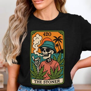 Könnte beinhalten: Schwarzes T-Shirt mit einem Tarotkarten-Design. Es zeigt ein Skelett mit Sonnenbrille und Fischerhut, das raucht. Die Karte trägt die Aufschrift "420" und "THE STONER" mit Cannabisblättern und einer Strandszene.