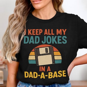 Könnte beinhalten: Schwarzes T-Shirt mit dem Text "I KEEP ALL MY DAD JOKES IN A DAD-A-BASE". Das Design zeigt eine Diskettengrafik vor einem Retro-Sonnenuntergang in Orange, Gelb, Türkis und Braun.
