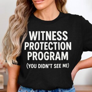 Könnte beinhalten: Schwarzes T-Shirt mit dem Text "WITNESS PROTECTION PROGRAM (YOU DIDN'T SEE ME)" in großen, weißen, fetten Buchstaben. Das Shirt hat einen Rundhalsausschnitt und scheint aus einem weichen Material zu sein.