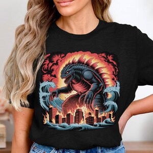 Könnte beinhalten: Schwarzes T-Shirt mit einer farbenfrohen Grafik eines Monsters, das eine Stadt zerstört. Das Monster ist dunkelblau und rot, mit einem feurigen Hintergrund und Meereswellen. Die Gebäude stehen in Flammen.