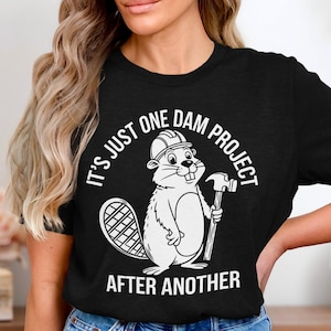 May include: Black t-shirt featuring a cartoon beaver wearing a hard hat and holding a hammer. The text on the shirt reads "IT'S JUST ONE DAM PROJECT AFTER ANOTHER" in a circular design around the beaver.