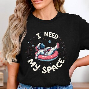 May include: Black t-shirt featuring a graphic of an astronaut floating in space on an inflatable ring. The text "I need my space" is printed on the shirt.