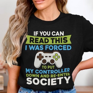May include: Black t-shirt with the text "IF YOU CAN READ THIS I WAS FORCED" and a controller graphic. The shirt's design includes the phrase "TO PUT MY CONTROLLER DOWN AND RE-ENTER SOCIETY."