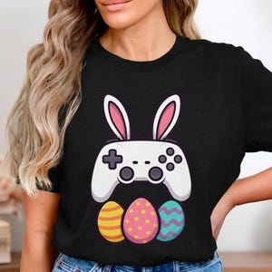 Könnte beinhalten: Schwarzes T-Shirt mit einem weißen Videospiel-Controller mit Hasenohren und drei bunten Ostereiern. Die Eier sind gelb mit orangefarbenen Streifen, rosa mit gelben Punkten und türkis mit lila Zickzack.