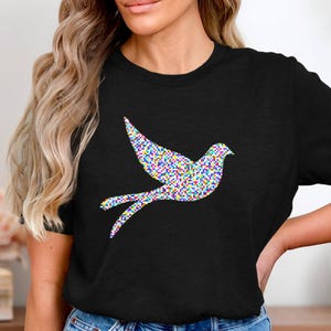 Könnte beinhalten: Schwarzes T-Shirt mit einem farbenfrohen, pixeligen Tauben-Design. Die Taube wird im Flug dargestellt, mit ausgebreiteten Flügeln und nachziehenden Schwanzfedern. Das Design verwendet ein Mosaik aus kleinen Quadraten in verschiedenen Farben.