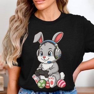 Könnte beinhalten: Schwarzes T-Shirt mit einem Cartoon-Hasen, der ein Headset trägt und einen Gamecontroller hält. Der Hase ist grau mit rosa und weißen Akzenten und sitzt neben Ostereiern. Das Design ist verspielt und farbenfroh.