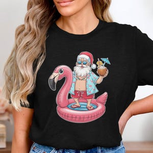 Puede incluir: Una camiseta negra con un gráfico de Santa Claus con una camisa hawaiana y gafas de sol, montando un flotador de flamenco rosa con una bebida de coco en la mano.