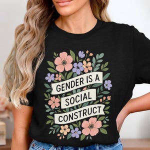 Könnte beinhalten: Schwarzes T-Shirt mit floralem Design und dem Text "GENDER IS A SOCIAL CONSTRUCT". Das Design umfasst rosa, lila und weiße Blumen mit grünen Blättern. Das T-Shirt hat einen Rundhalsausschnitt.