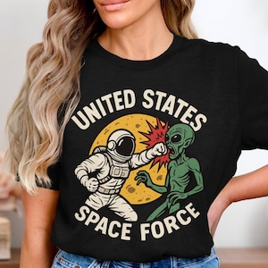 Puede incluir: Camiseta negra con un dibujo animado de un astronauta golpeando a un extraterrestre. El diseño incluye el texto "UNITED STATES SPACE FORCE" en una disposición circular alrededor de las figuras, sobre un fondo amarillo.