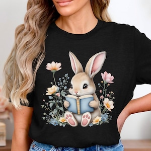 May include: A black t-shirt with a graphic of a brown bunny rabbit sitting in a field of flowers and reading a blue book. The bunny has pink ears and a white nose.