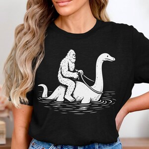Könnte beinhalten: Schwarzes T-Shirt mit einer weißen Grafik von Bigfoot, der das Ungeheuer von Loch Ness reitet. Die Illustration zeigt die Kreaturen im Wasser in einem einfachen Linienstil. Es ist ein lässiges Kleidungsstück.